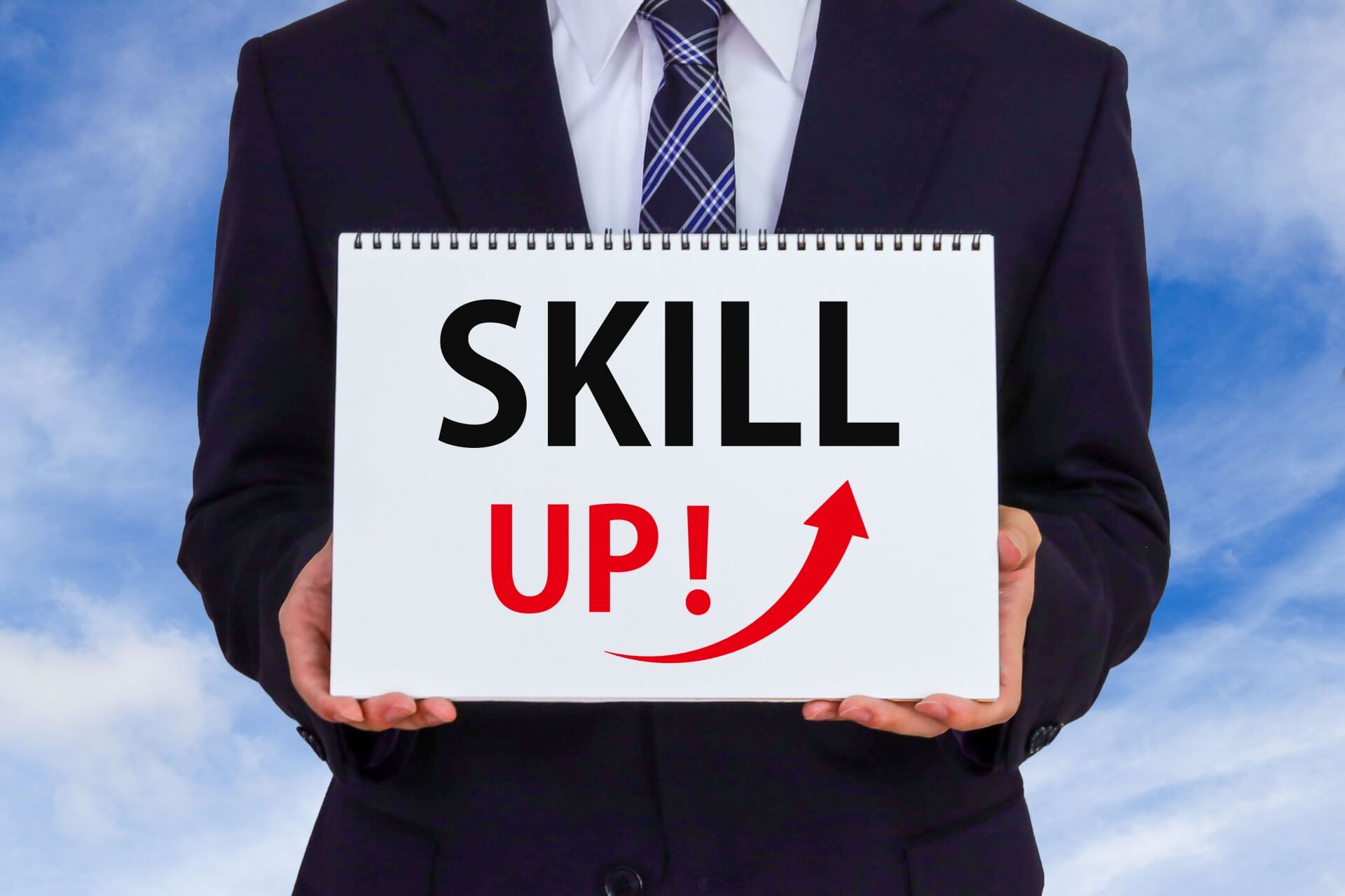 「SKILL UP！」と書かれたボードを胸元で持つスーツ姿の男性。自律的なスキルアップや自己啓発を促す、社員教育のイメージ。