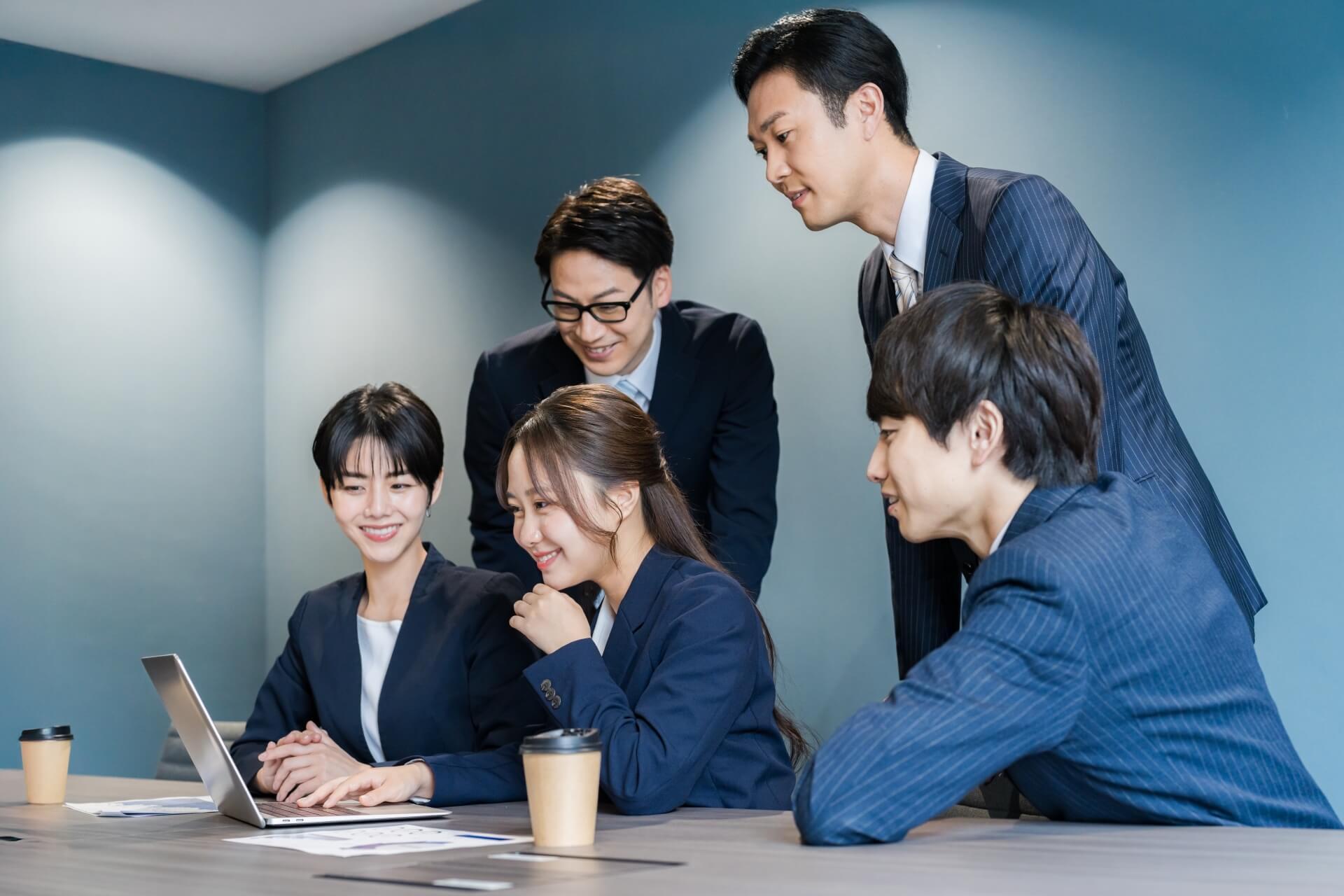 ノートパソコンを囲み、笑顔で打ち合わせをする5人のビジネスチーム。集合的効力感が高い組織の協力的な様子。