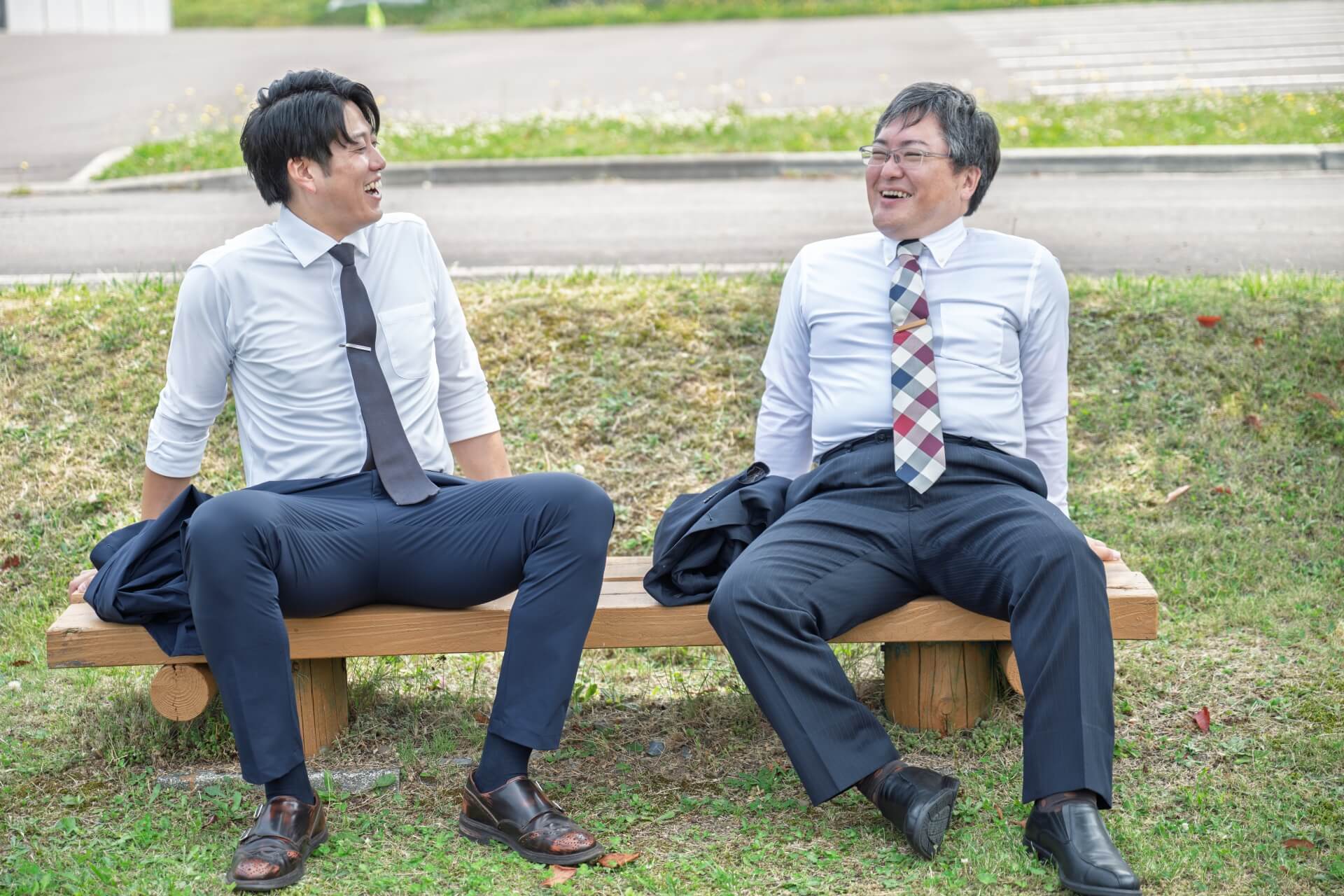 屋外のベンチに座り、リラックスした表情で本音を語り合い笑い合う2人の男性ビジネスパーソン