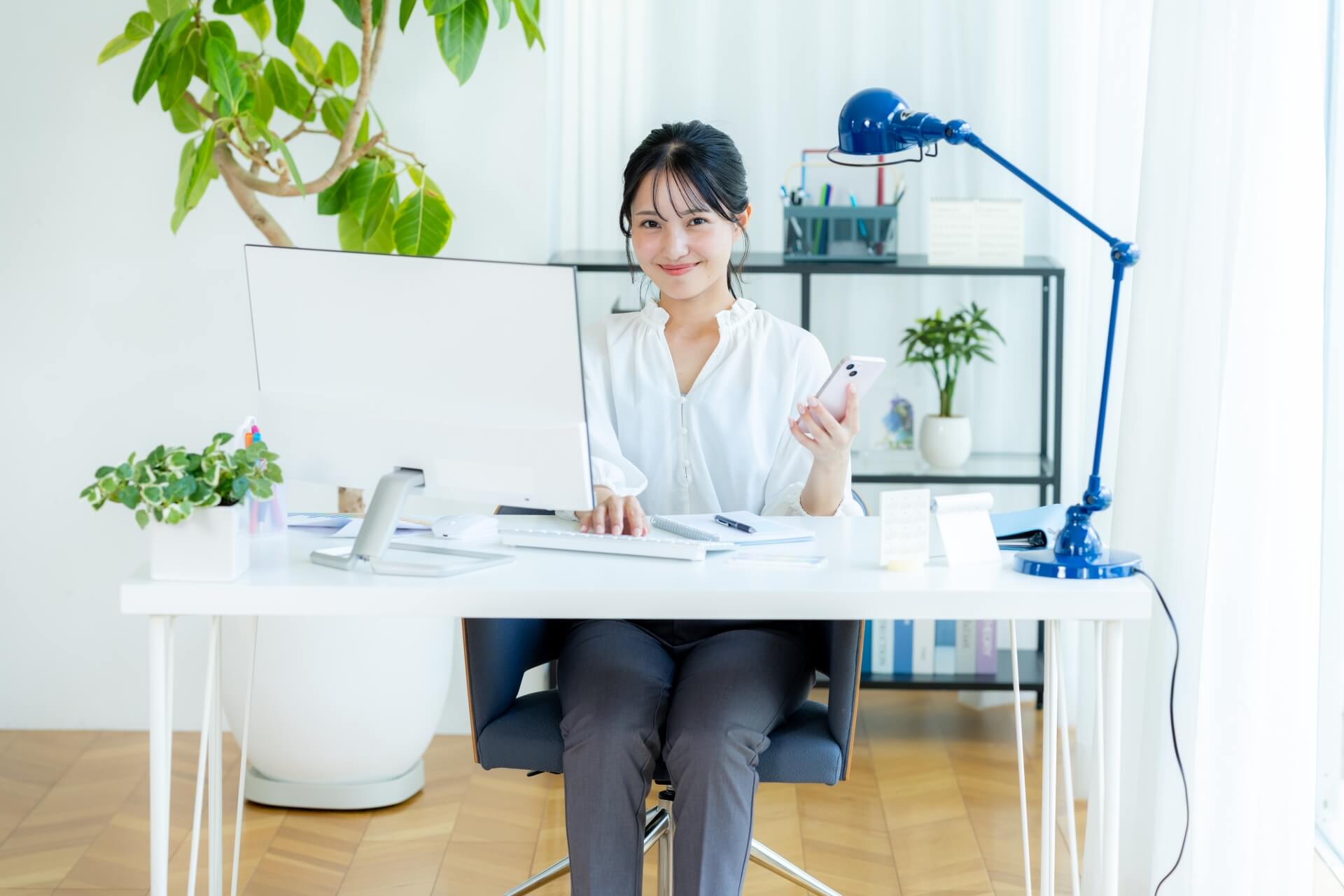 デスクで自律的に業務を進め、スマホで情報を確認する笑顔の女性社員