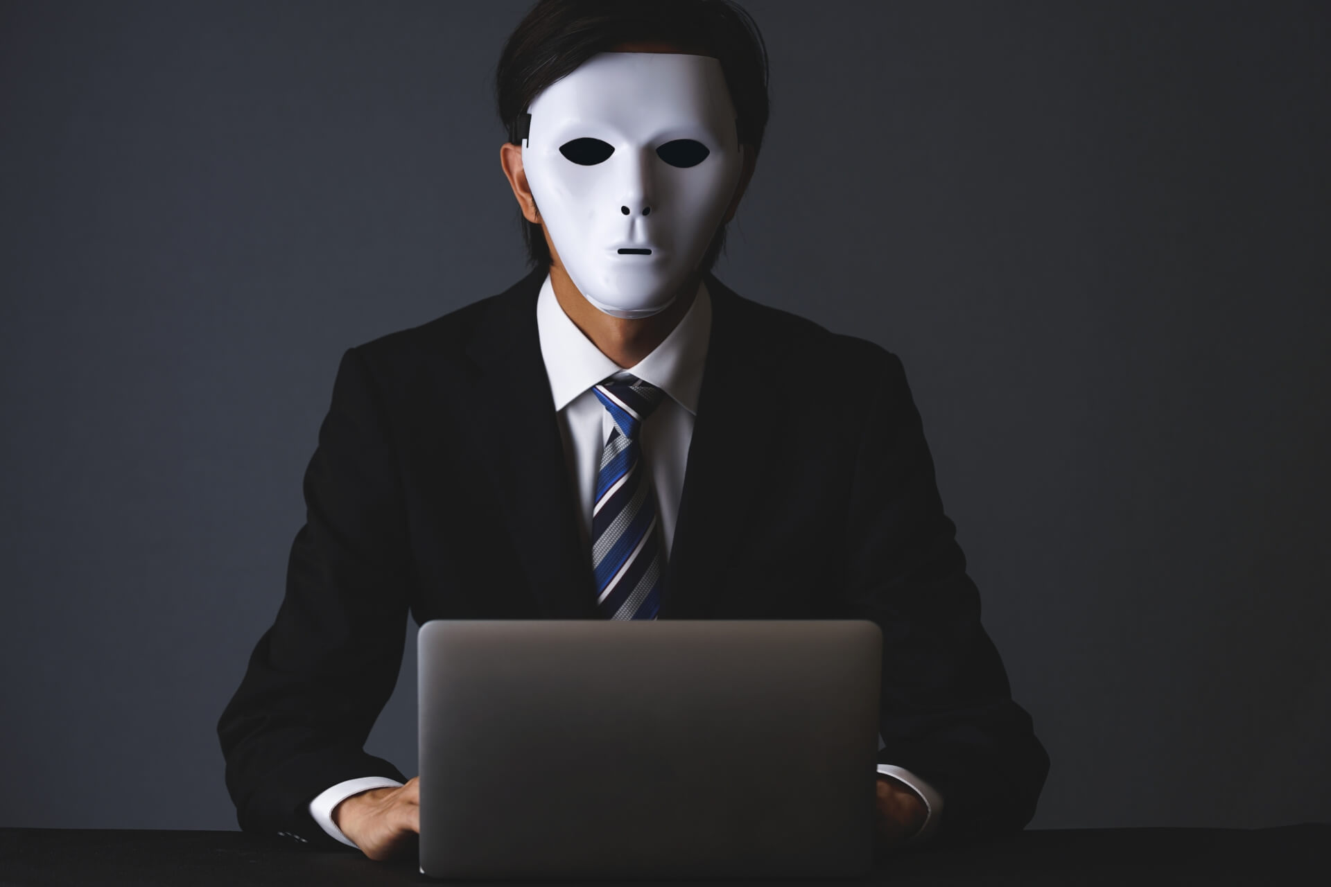白い仮面をつけてパソコンに向かうスーツ姿の男性。静かな退職や組織への諦め、本音が見えない従業員の状態を表現したイメージ。