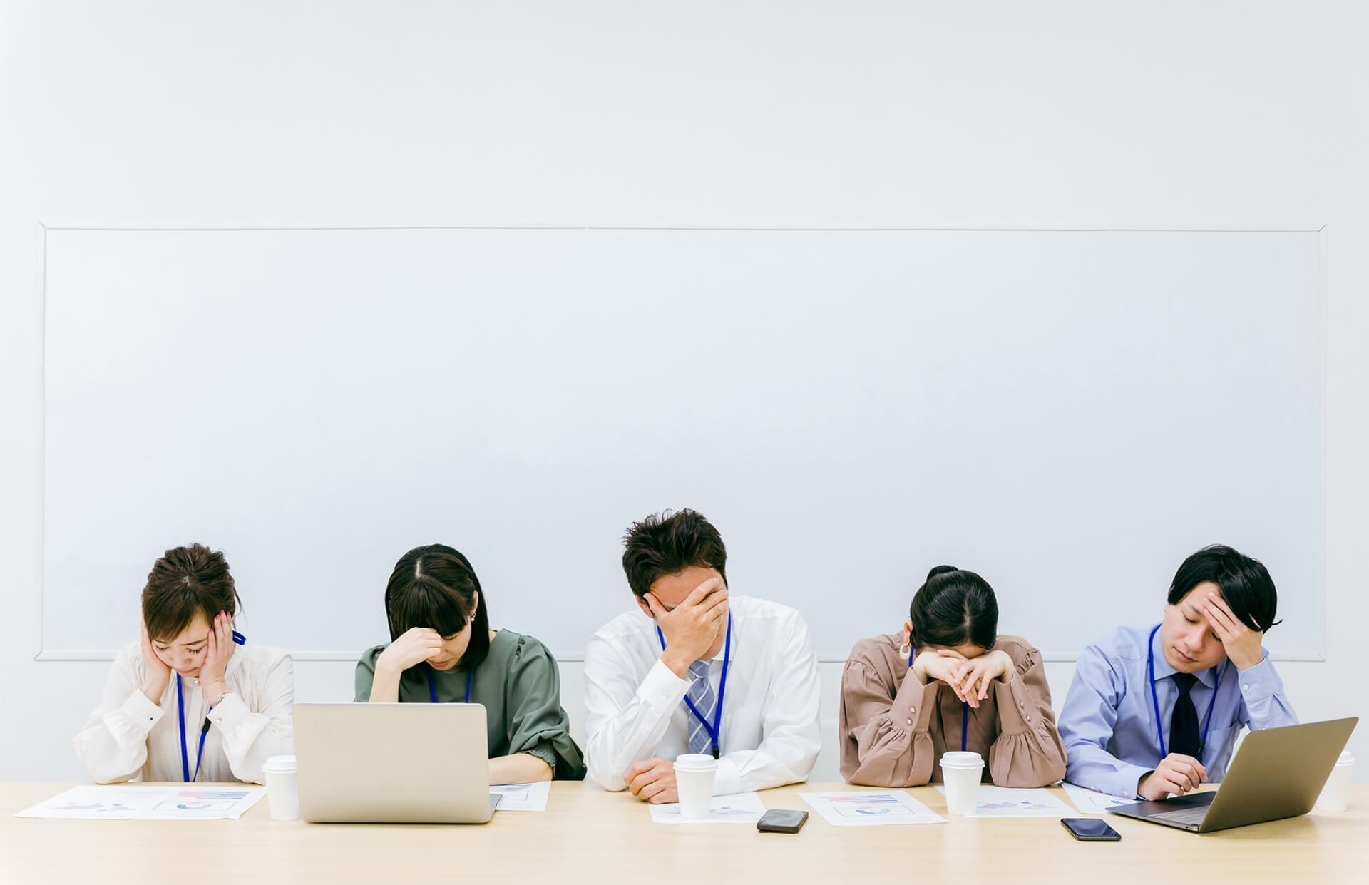 会議室のデスクで一列に並び、全員が頭を抱えたり俯いたりして意欲を失っているビジネスパーソンのグループ。職場におけるモチベーション低下の伝染や、重苦しい組織の雰囲気を表現したイメージ
